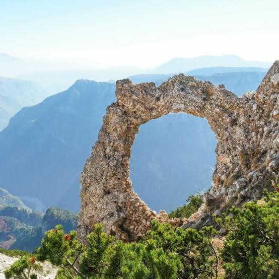 Natural stone arch on a karst ridge above hazy Herzegovina ranges, dwarf pine in front