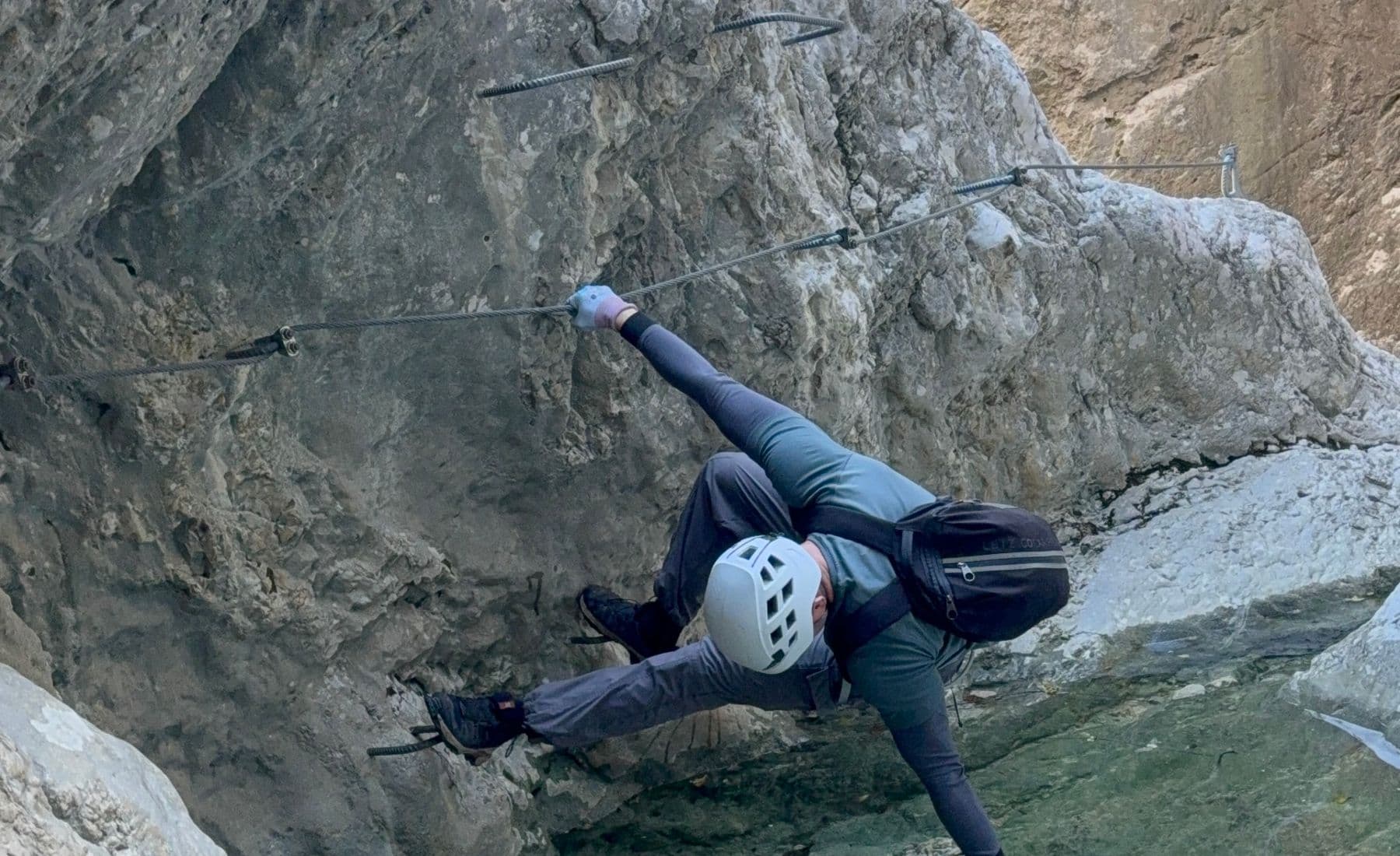Climber traversing the cable above a green pool in the Podružje canyon on the Ilirski Put route