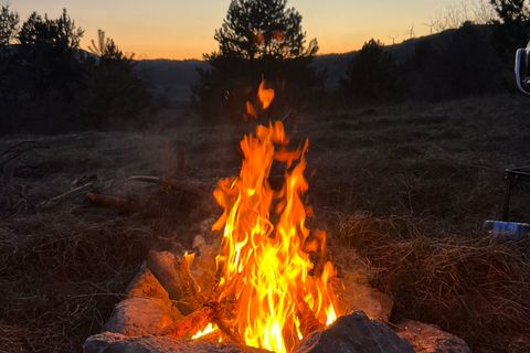 Campfire burning inside a ring of stones at dusk, pines and low hills behind the camp