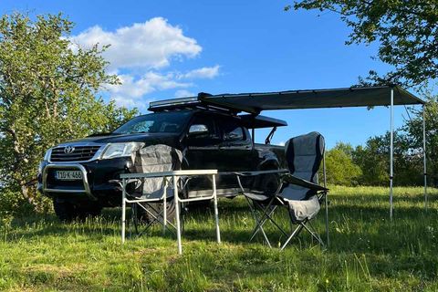Pickup with a roof rack and awning beside a camp table and chair on a grassy meadow