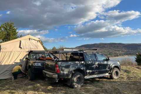Two 4x4s and a large tent pitched on a grassy bluff above a lake, wooded hills beyond